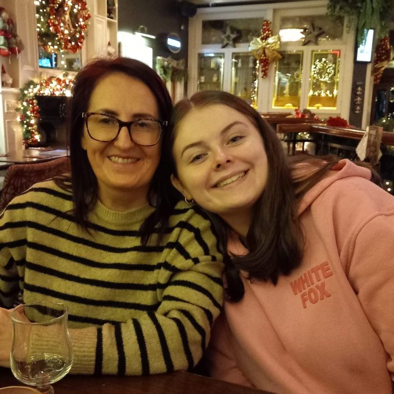 Timeless Stitches Two smiling women with glasses, sitting at a table decorated for the holidays.